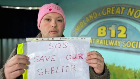 A man standing inside the Sheringham Bus Shelter which has a painted train mural on the inside of it. He is looking directly at the camera and is wearing a pink beanie, with a yellow fluorescent hi-vis vest. He is holding a paper sign which says SOS SAVE OUR SHELTER.