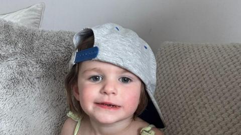 Three-year-old Teraysa looks into the camera sat on a sofa with a baseball cap on sideways.