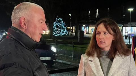 Jim Cunningham and Lola McEvoy standing outside on the street, speaking to each other. There is a Christmas tree with blue lights in the background. It is dark and street lights are on. Mr Cunningham has grey hair and is wearing a thick black coat. McEvoy is wearing a light grey coat and has brown hair. She is also wearing a grey turtleneck.