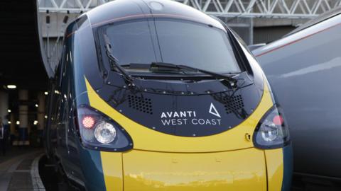 Close up of the front of a yellow and navy Avanti West Coast train at a platform at a rail station.