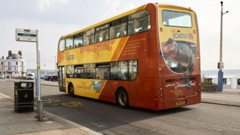 An image of a yellow, orange and red bus on a road with a bus stop on one side and the seaside on the other.