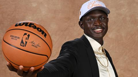 Khaman Maluach wears a Phoenix Suns baseball cap and a suit as he poses with an NBA-branded basketball