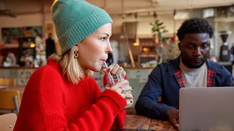 A young woman wearing a red woolly jumper and teal woolly hat sits at a wooden table in a cafe sipping a drink. A man wearing a blue coat uses a laptop to her right.