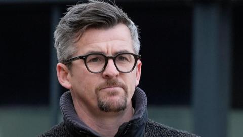 Joey Barton with grey hair and brown beard and black glasses wearing a grey hoodie.