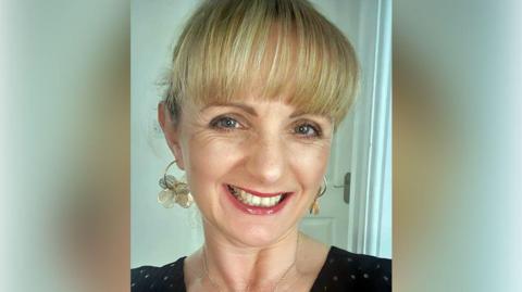 Gail Hardy with a long blond fringe and the rest of her hair pinned back standing in front of a white door in a house. She is wearing a black sparkly top and gold and white hooped earrings with flowers. She is smiling.