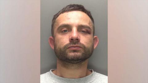 A police head and shoulders shot of a man with short brown hair looking into the camera with a serious expression. He has brown stubble and a moustache and is wearing a grey top. One of his eyes appears drooped and bruised.