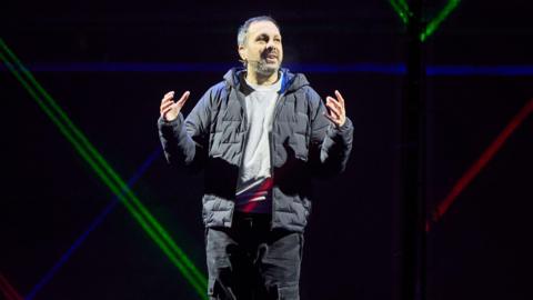 Steven Frayn stands on stage with his hands gesturing he is a man with short brown-grey hair. He is wearing black cargo trousers, a grey jumper and a black puffer jacket. Behind him are coloured lights.