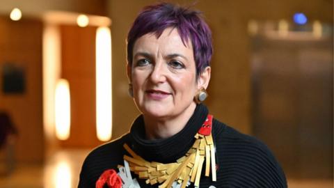 Angela Constance, who has short hair dyed purple, in the Scottish parliament. She is wearing a black turtle-neck jumper with a gold and red decorative piece clipped on.