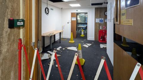 A red and white barrier preventing access to a small room. The room has a damaged roof and chunks of ceiling tiles are lying on the carpeted floor alongside yellow warning cones.