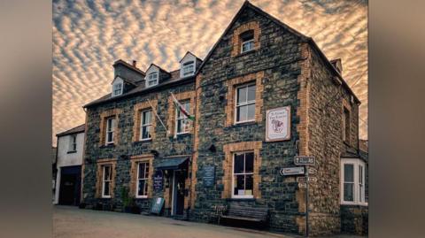 Llun o dafarn wedi'i adeiladu allan o gerrig gydag awyr cymylog tu ôl iddo.