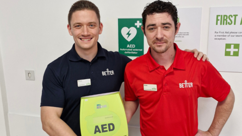 The image shows two men holding a neon Automated External Defibrillator in their hands while posing for a picture. The man on the left is wearing a black shirt and the man on the left a red one, both with "Better" written on the left ha-hand-side of their chests. The wall behind them is white with First Aid signs on it. A light switch can also be seen on the left-hand-side of the wall.