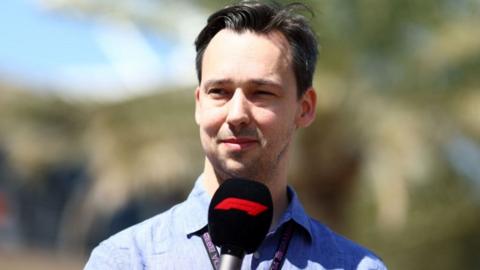 Alex Jacques looks off camera. He has black swept-back hair and is wearing a blue shirt. A lanyard is around his neck and his is holding a black and red F1-branded microphone.
