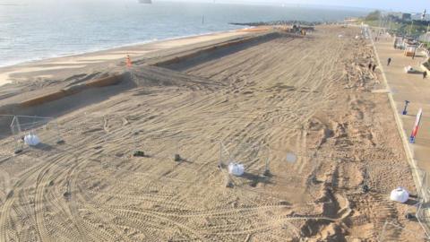 Beach with golden shingle and promenade to the right and sea to the left - fencing can be seen creating a rectangular section in the centre.