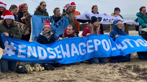 A group of people are kneeling and standing together for a photograph on the sandy beach with the sea behind them on a grey day. Most of them are wearing red and white Christmas hats are are wrapped up in winter coats and scarves. The largest banner reads: #END SEWAGE POLLUTION.