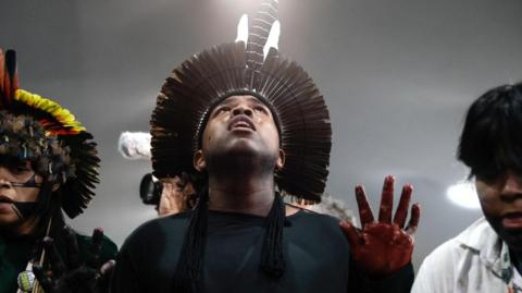 Indigenous people at a protest. Man in centre wears a headpiece made of features and red dye or ink on fingers