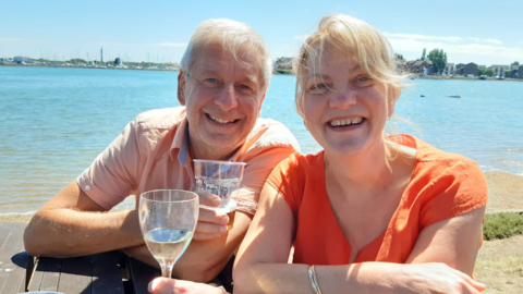 Chris and Ruth Stone-Houghton sit on a picnic bench, smiling into the camera. They are holding alcoholic drinks, and behind them is a body of water