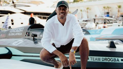 Didier Drogba, wearing a black gap, loose white shirt and black shirts, crouches down as he poses in front of a black powerboat with light blue detailing which has the slogan Team Drogba on its prow. In a marina setting, several other boats, including one very large superyacht, can be seen in the background