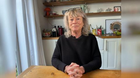 A blonde woman sits at a wooden table in her home.