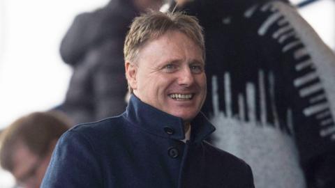 Billy Hewitson, who has spiky blond hair, is wearing a dark jacket with the collar turned up. He is smiling.