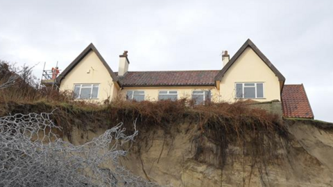 A view from the bottom of a cliff looking up toward a house that can be seen towering over. It is a yellow house with several windows and two chimneys. Only the top storey of the house can be seen. Metal wires can be seen toward the bottom left of the image as part of sea defences.