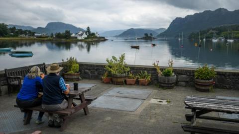 A woman and a man, who is wearing a wide brimmed hat, sit at a picnic bench drinking coffees and looking out over the bay at Plockton in Wester Ross.