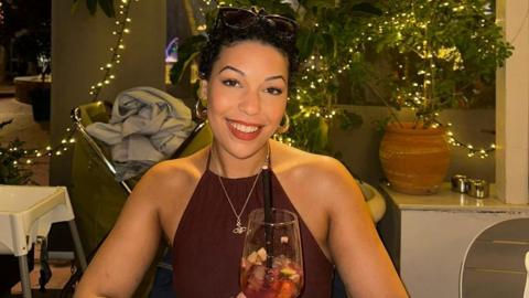 Sacha Gomez, sitting in a bar, looking at the camera and smiling, she has short dark hair, with sunglasses on her head. She has earrings, a necklace and a burgundy top. There are decorations, and lights behind her.