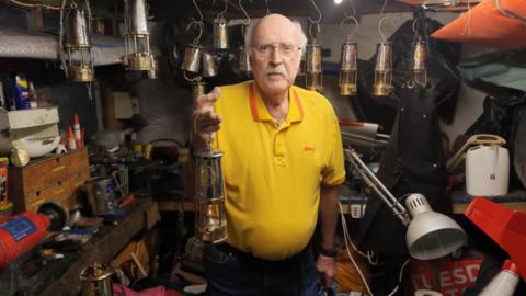 A man with short, white hair, a white moustache, glasses, a yellow polo shirt and dark trousers is holding a miners lamp. He is standing in a shed filled with other miners lamps and tools.
