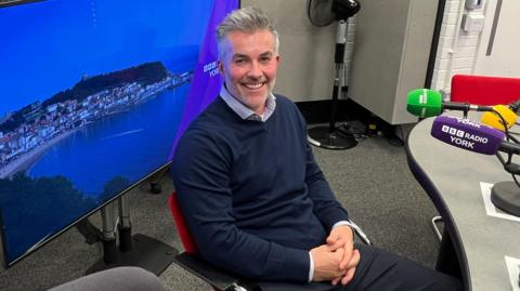 Labour mayor of York and North Yorkshire, David Skaith, is sitting in the BBC Radio York hotseat, waiting to be interviewed by presenter Joanita Musisi. He is smiling at the camera, wearing a blue jumper and an open-neck shirt and has his hands clasped in front of him