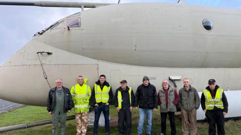 A group of men standing in front of a vintage plane on an airfield