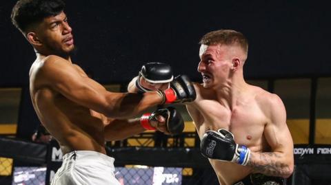 Two men are in the boxing ring, one is ethnically diverse the other is white. The white fighter has the upper hand and has gritted teeth. His opponent has a dazed expression. You can see the veins in the head of the white fighter