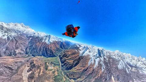 A man in a red suit, helmet and goggles flying over snow-capped mountains