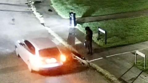 A CCTV still of a car driving past a man, dressed entirely in black, standing on the edge of a road waiting to cross. The man has just come from a footpath which goes between two patches of grass. The car's licence plate is not visible.