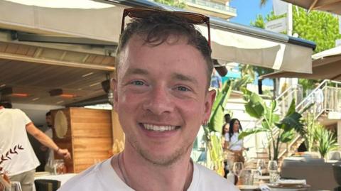 Calum sits in a sunny, outdoor restaurant. He has sunglasses on his head and is smiling. He has blue eyes and two small silver hoop earrings in his left ear.