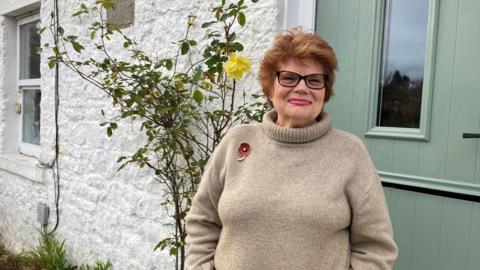 Vanessa Corby is standing in front of her door. She is wearing a brown jumper with a poppy pinned to her right shoulder. She has short, styled reddish brown hair and black-rimmed glasses and smiles to the camera. Behind her is a white wall with a yellow rose bush growing up it. There is a green panelled door with a central window over her left shoulder.