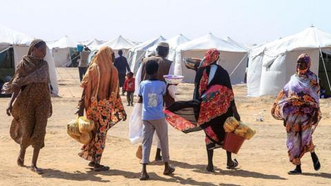 Four women, a boy and a man walking on sandy ground carrying food. In the background are white aid tents.