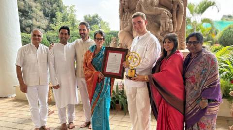 A group of people in south Asian attire are smiling at the camera while the two people in the middle are holding awards