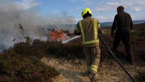 A firefighter in full protective gear, including a high-visibility helmet and suit, sprays water onto a wildfire burning through dry vegetation. Thick smoke and flames rise in the background. Another person in dark clothing stands nearby, watching. The scene takes place in a rural, hilly area with sparse greenery.