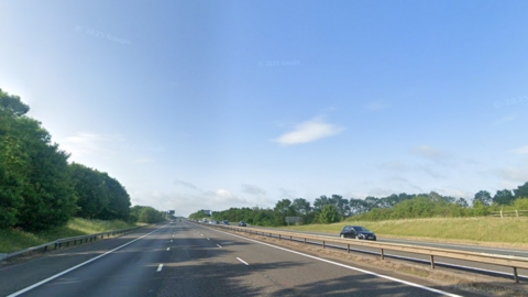 A motorway with green verges on either side. The sky is blue and there are trees on each side of the road. There is a single black car on the opposite carriageway.