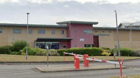 Exterior of the prison, a red and cream wide building, with red and white barriers in front of it.