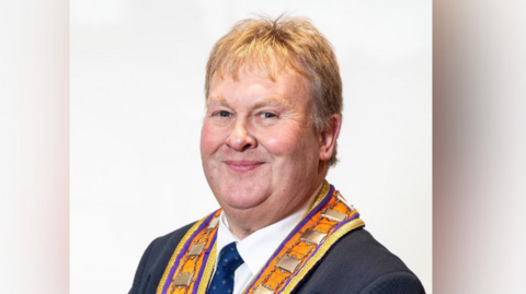 A professional photograph of Harold Henning. He is standing in front of a white background. He is an older man, with medium-length grey hair. He is wearing a Orange Order sash over the top of his dark coloured suit jacket, blue tie and white shirt. He is smiling at the camera.