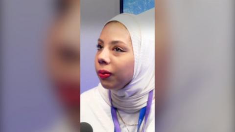 A WOMAN LOOKS AWAY FROM THE CAMERA. SHE WEARS A WHITE HEADSCARF