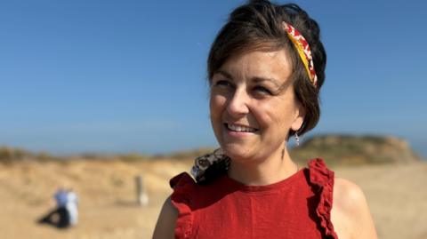 A smiling Sarah Wheldon head and shoulders. She is standing on a beach on a sunny day wearing a red high-neck sleeveless top with a frill trim around the arms. She has brown hair that reaches her neck and a red, white and yellow tie-died scarf tied as a headband.