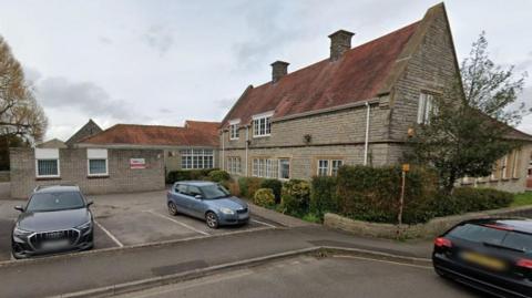 A street view image of a stone building with a car park to the left. There are two cars in the car park. There is a tree and a hedge at the front of the building