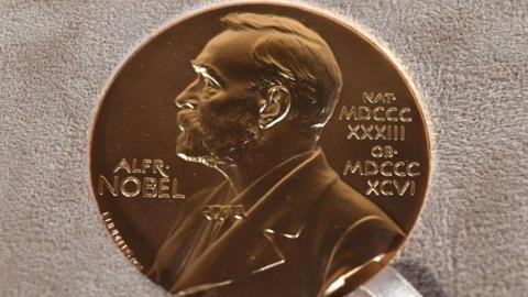 A gold medal lying on a cloth with the etched outline of a man's head and torso with the words Alfred Nobel