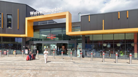 A metal, yellow bar runs along the front of the station a few metres off the ground and goes higher and forms a square shape above the glass door entrance to the station. Bollards are across the entrance and glass windows either side as some passengers are coming out.