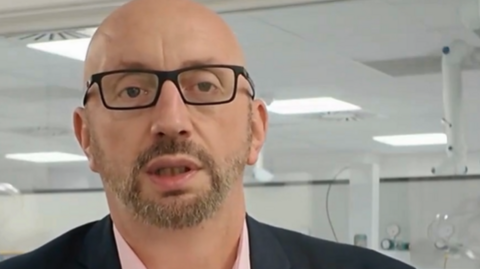 A close up image of a bald white man with a beard and black-rimmed glasses looking at a camera.