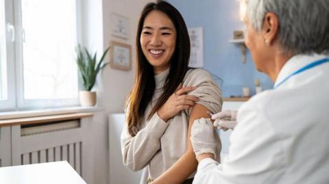 A woman receives a flu vaccine from a doctor. The woman is holding her sleeve up and smiling and the woman giving the vaccine is wearing a white coat. The room has blue and white walls and they are sitting near a window with a plant on the windowsill.