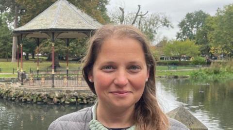 A woman with brown hair and a grey top is looking at the camera. A bandstand, which is next to water, is behind her on the photo.