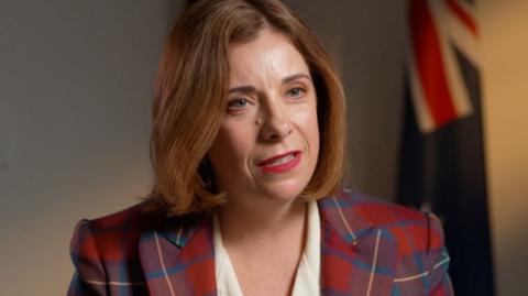 A woman in a tartan jacket and white dress speaks. Several flags can be seen in the background