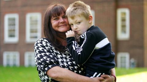 A woman who is holding her son in her arms. They are both wearing black and white, she has long dark hair and he has short blonde hair. A building and grass are blurred in the background.